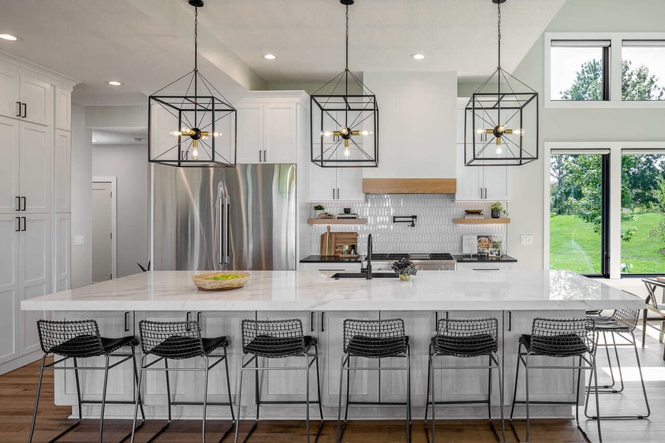 Large marble island in a kitchen surrounded by steel wired seats, pendant lights and large windows